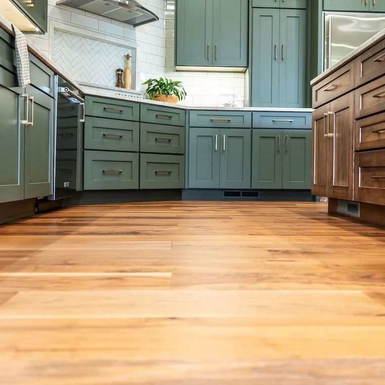 Modern kitchen featuring warm laminate flooring, green and brown shaker cabinets, and stainless steel appliances with a tiled backsplash